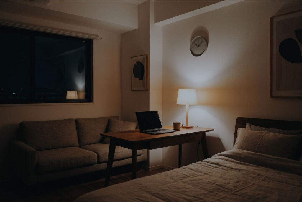 静かな夜の部屋で、間接照明の温かい光がデスクやベッドを優しく照らす落ち着いた空間。