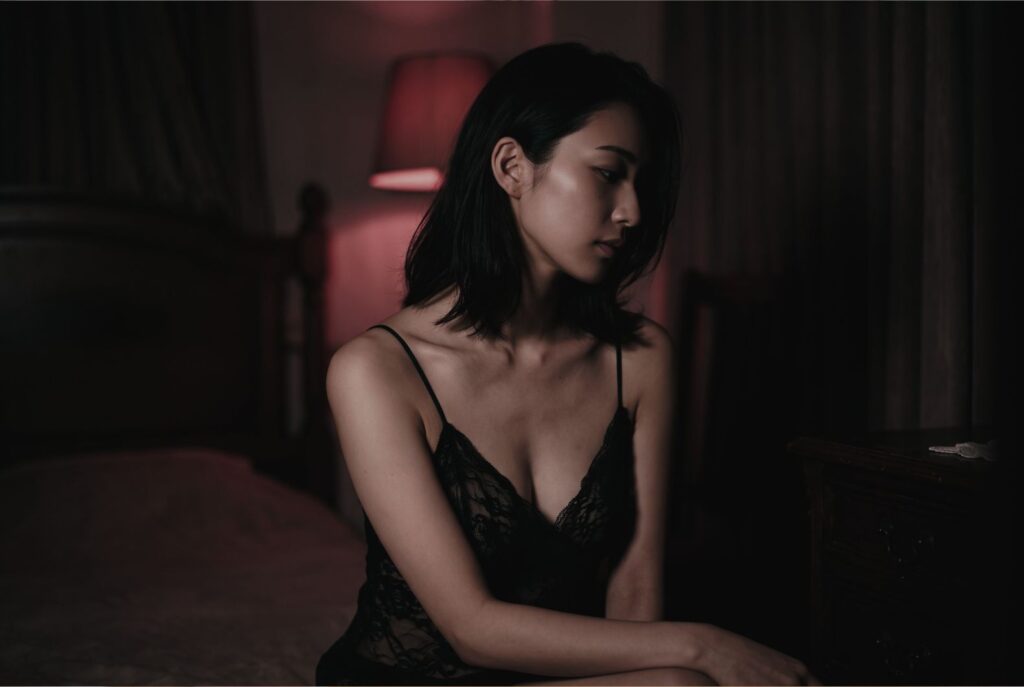 薄暗く赤い間接照明が灯る部屋で女性が伏し目がちに佇む様子。